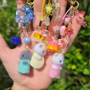 May include: A collection of handmade keychains featuring miniature plush animal figures in pastel colors. Each keychain has a gold-colored clasp and is adorned with beads, charms, and translucent star and geometric shapes. The keychains are held in a hand.