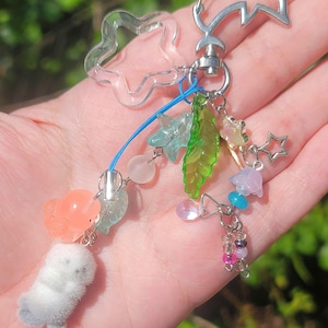 May include: A whimsical keychain with a dolphin-shaped clasp and a clear star-shaped charm. It features a variety of colorful charms, including a fuzzy white seal, translucent leaves, stars, and beads in shades of pink, blue, and green.