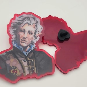 Puede incluir: Un pin de esmalte rojo con una ilustración detallada de un hombre con cabello plateado y cuello blanco. El pin tiene un cierre de metal negro.