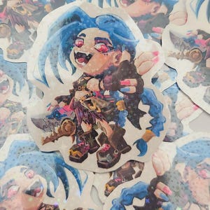 May include: A stack of holographic stickers featuring a cartoon character with blue hair, red eyes, and a toothy grin. The character is wearing a black and white outfit and holding a weapon. The stickers have a sparkly, iridescent finish.