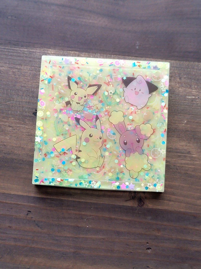 May include: Square resin coaster with a light yellow background, featuring cartoon characters and colorful confetti. The coaster showcases Pikachu, Pichu, and other characters. The design includes glitter and small decorative elements.