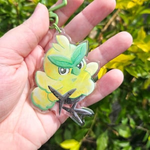 May include: A clear acrylic keychain featuring a yellow, bird-like creature with green leaf-shaped eyebrows and dark brown, pointed feet. The character has a rounded, fluffy body and is attached to a light green plastic lobster clasp hook.