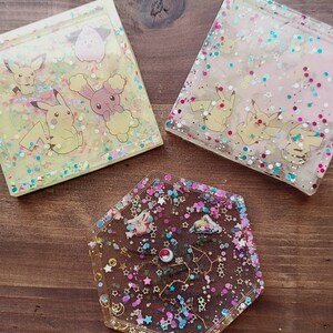 May include: Three resin coasters with colorful designs. Two square coasters feature Pikachu and other cartoon characters with glitter. The third coaster is a hexagon shape with a constellation design and glitter.
