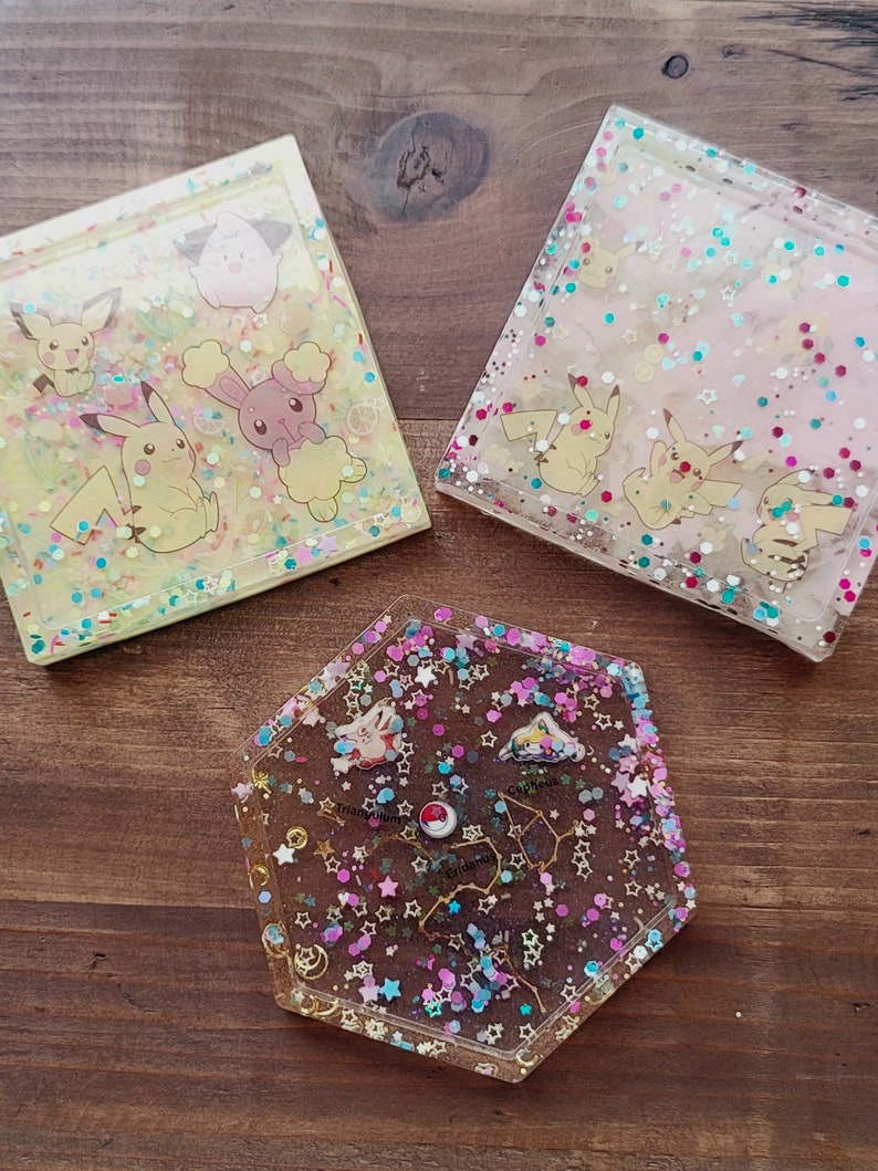 May include: Three resin coasters with colorful designs. Two square coasters feature Pikachu and other cartoon characters with glitter. The third coaster is a hexagon shape with a constellation design and glitter.