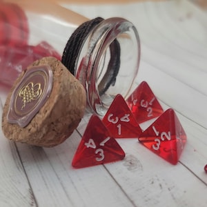May include: A clear glass bottle with a red tint, a cork stopper, and several red, triangular dice with white numbers. The cork has a gold emblem. The dice are scattered on a light-colored wooden surface.
