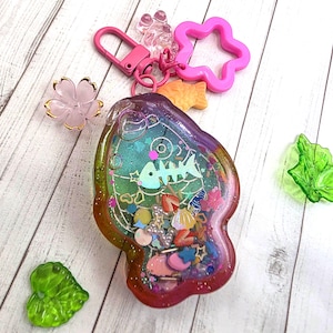 May include: A fish-shaped resin shaker keychain featuring a gradient of purple, green, and orange. The interior contains a holographic fish skeleton, small colorful beads, stars, and shell charms. It includes a pink lobster clasp and a star charm.