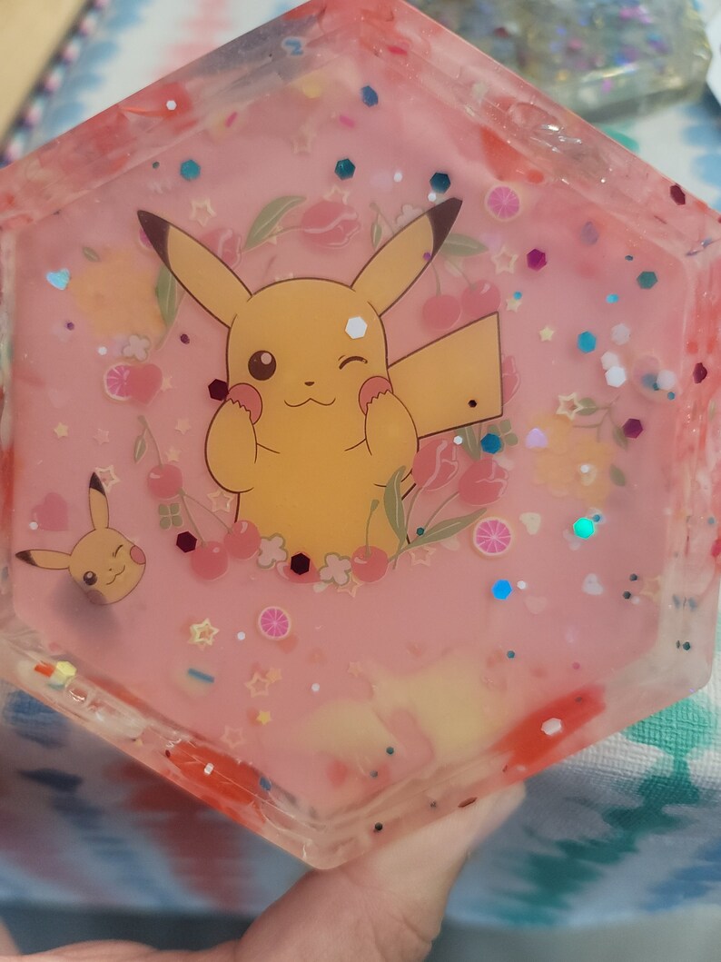 May include: A hexagonal resin tray with a pink background, featuring a cheerful Pikachu design surrounded by flowers, cherries, and glitter. The tray has a clear, glossy finish and is suitable for holding small items.