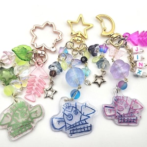 May include: A selection of handmade keychains with diverse charms. Each keychain showcases a unique design, such as stars, fish bones, and abstract figures. The keychains are decorated with colourful beads and small charms, with a gold-tone clasp.