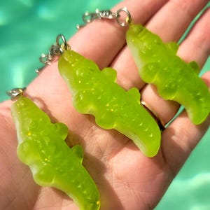 Op de afbeelding: Drie groene sleutelhangers in de vorm van een alligator met zilverkleurige hardware. De sleutelhangers zijn doorschijnend groen met een glitterachtig uiterlijk. De achtergrond is een wazige turquoise kleur.