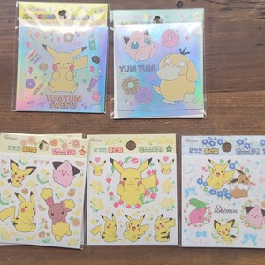 May include: A collection of Pokemon stickers in clear plastic packaging. The stickers feature Pikachu and other characters with the words "Yum Yum Sweets" and "Pokemon". The stickers have a holographic background.