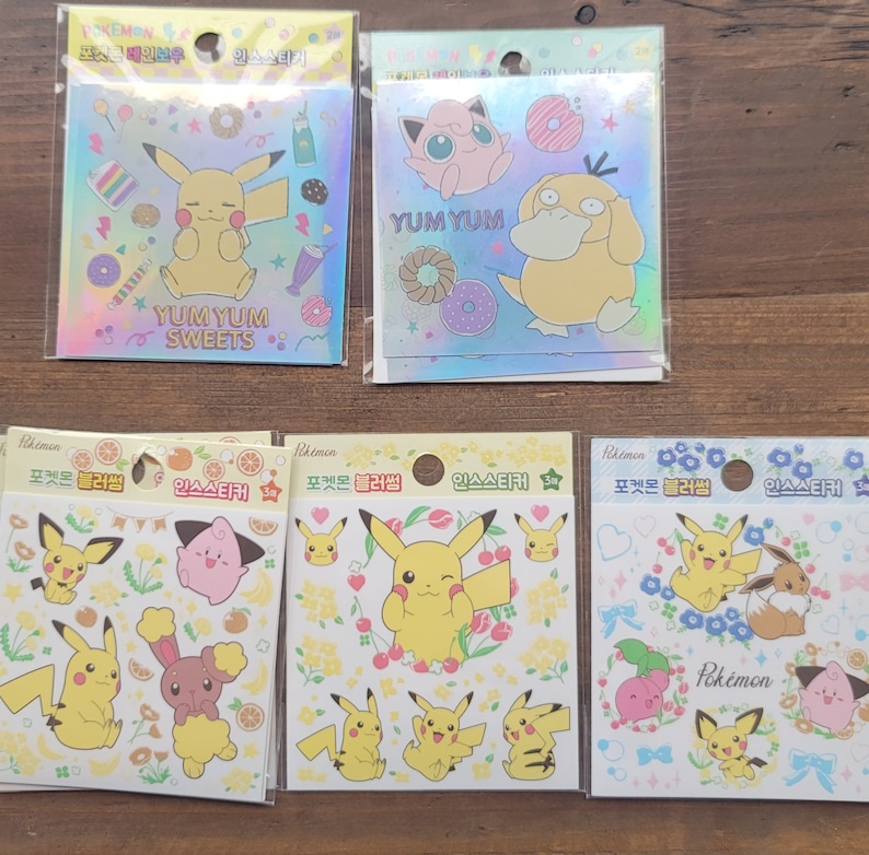 May include: A collection of Pokemon stickers in clear plastic packaging. The stickers feature Pikachu and other characters with the words "Yum Yum Sweets" and "Pokemon". The stickers have a holographic background.