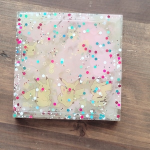 May include: Square resin coaster with a clear border, featuring a pink and white background with glitter and Pikachu characters. The coaster has a glossy finish and is set on a wooden surface.