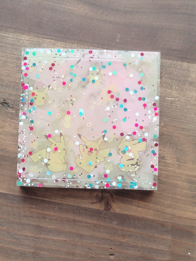 May include: Square resin coaster with a clear border, featuring a pink and white background with glitter and Pikachu characters. The coaster has a glossy finish and is set on a wooden surface.