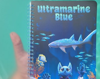 May include: A blue spiral bound notebook with a cover featuring a cartoon cat looking up at a shark swimming in a blue ocean with coral and fish. The cover text reads "Ultramarine Blue".