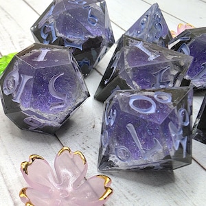 May include: A set of polyhedral dice featuring a translucent purple core with a jagged, crystalline texture encased in a clear, dark resin shell. The numbers are engraved in a light blue font. A small pink flower-shaped object is in the foreground.