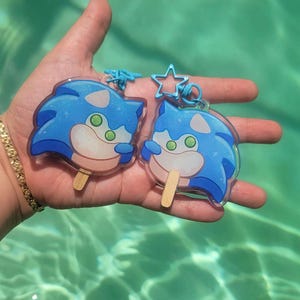 May include: Two blue and white Sonic the Hedgehog-themed popsicle keychains. Each keychain features a cartoon character design with green eyes and a wooden popsicle stick. The keychains have blue star-shaped and swirl-shaped clasps.