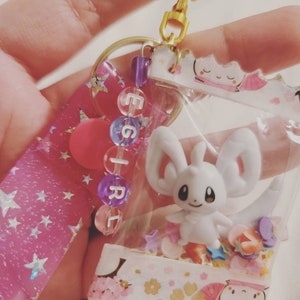 Può includere: Un portachiavi in plastica trasparente con una piccola figurina di Eevee bianca all'interno. Il portachiavi è pieno di glitter e perline colorate, e ha un bordo con motivo rosa e bianco. Il portachiavi ha anche un nastro rosa e bianco con stelle argentate.