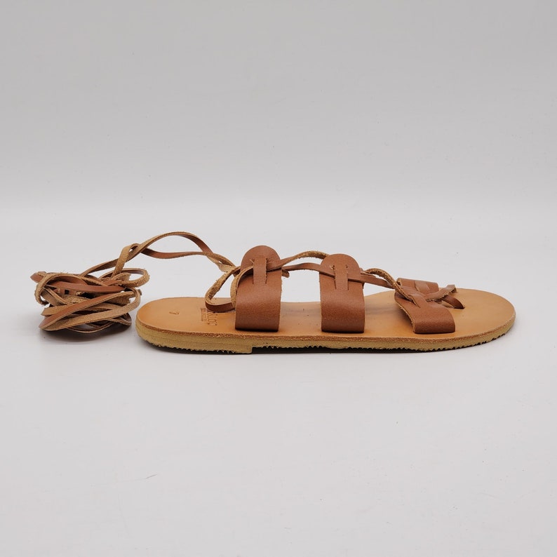 May include: Brown leather sandals with a lace-up design. The sandals have a flat sole and are tied around the ankle.