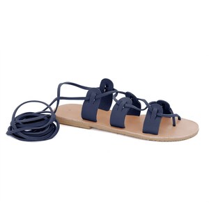 May include: Navy blue leather sandals with a lace-up design. The laces are tied around the ankle and the sandals have a flat sole.