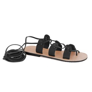 May include: Black leather sandals with a lace-up design. The laces are tied around the ankle and the sandals have a flat sole.