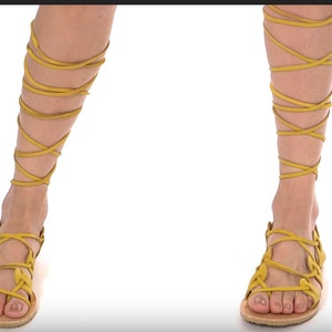 May include: A pair of yellow leather sandals with laces that wrap up the legs. The sandals have a simple design with a flat sole.