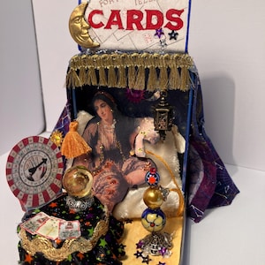May include: A handcrafted fortune-telling box with the text "Madam Morrow's Fortune Telling Cards." The box features a gold crescent moon, a miniature fortune teller image, a crystal ball, and a deck of cards. The box is decorated with gold fringe and star accents.