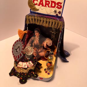 May include: A miniature fortune-telling diorama featuring a sign that reads "Madam Morrow's Fortune Telling Cards." The scene includes a fortune teller image, a crystal ball, and decorative elements. The diorama is housed in a small, decorated box.