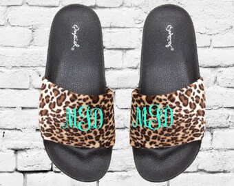 Op de afbeelding: Een paar zwarte slippers met een luipaardprint band. De band heeft een bruine en bruine dierenprint en een turquoise monogram. De merknaam "Quupid" staat op het voetbed.