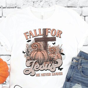 Puede incluir: Camiseta blanca con un diseño gráfico de una cruz con calabazas y mariposas. El texto "Fall for Jesus" y "He Never Leaves" está impreso en la camiseta.