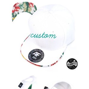 May include: White baseball cap with a floral print brim and a black and white logo that says "Academy Fits" on the side. The cap has a white strap with a black buckle and the text "custom" is embroidered on the front.