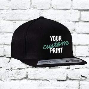 以下が含まれることがあります： 白いつばが付いた黒いベースボールキャップ。ターコイズグリーンの文字で「YOUR custom PRINT」と書いてあります。