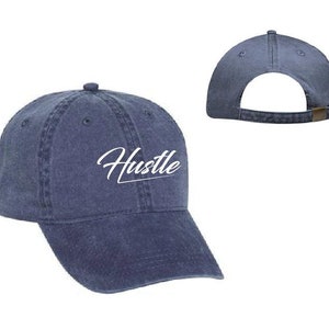 Op de afbeelding: Een marineblauwe baseballpet met het woord "Hustle" in wit geborduurd op de voorkant.