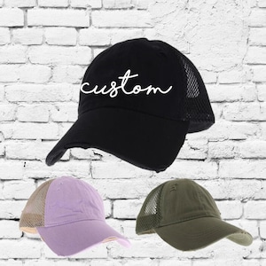 May include: Three distressed baseball caps with mesh back panels. The black cap has the word "custom" embroidered in white. The other two caps are lavender and olive green.