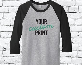 Può includere: Una maglietta raglan grigia con maniche nere. La maglietta presenta la scritta "YOUR custom PRINT" in nero e verde acqua. La maglietta è mostrata contro un muro di mattoni bianchi.