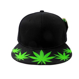May include: Black baseball cap with a flat brim. The brim features a repeating pattern of bright green cannabis leaves. The cap has a structured crown and a small, round button on top. A green and gold tag is attached to the side.