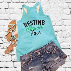 Puede incluir: Una camiseta sin mangas azul claro con las palabras "RESTING Beach Face" en negro y verde, combinada con shorts vaqueros y sandalias azul claro. Los shorts tienen detalles desgastados.