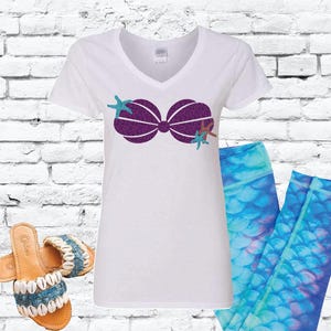 May include: White t-shirt with a purple glitter seashell design and a turquoise starfish. The shirt is paired with blue leggings with a mermaid scale pattern.