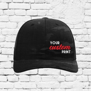 Puede incluir: Gorra de béisbol negra de camuflaje con parte trasera de malla y visera curva. La gorra tiene el texto "YOUR custom PRINT" en letras blancas y rojas. La gorra está sobre una pared de ladrillos blancos.