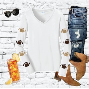 May include: White long-sleeve shirt with a V-neck and gold glitter footballs on the sleeves. The shirt is paired with blue jeans and brown ankle boots.