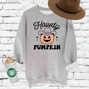 Puede incluir: Una sudadera gris con el texto "Howdy Pumpkin" en negro. Una calabaza de dibujos animados con un sombrero de vaquero está en el centro del texto. La sudadera está sobre un fondo de ladrillos blancos.