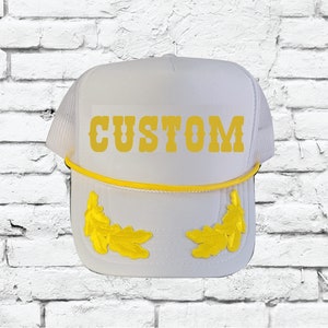May include: White trucker hat with a mesh back and a yellow rope detail. The word "CUSTOM" is printed in yellow on the front. Yellow leaf designs are embroidered on the front of the hat.