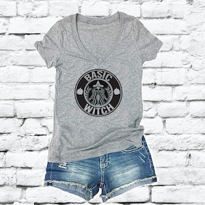 May include: A gray V-neck t-shirt with a black and white graphic that reads "BASIC WITCH" in a circular design. The design includes a witch, pumpkins, and a coffee cup logo. The shirt is paired with denim shorts.