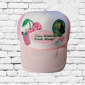 May include: Pink and white trucker hat with embroidered patches. Patches include cherries, a cocktail, a toucan, and hearts. The hat features the text "Save Water Drink Margs" in green script.