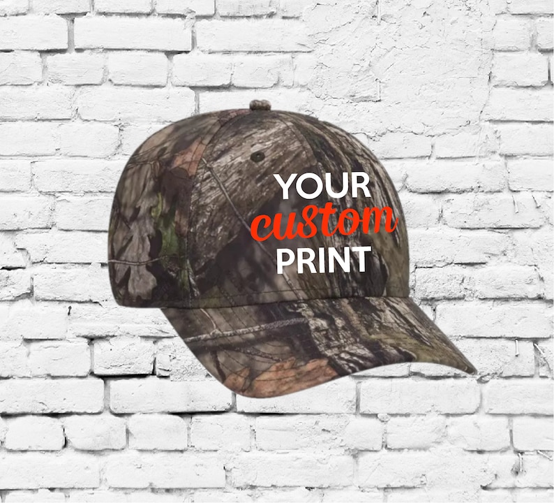 Op de afbeelding: Een camouflage baseballpet met de tekst "YOUR custom PRINT" in witte letters met een oranje omtrek.