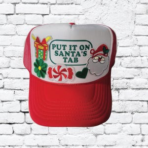 Peut inclure: Casquette de camionneur rouge et blanche avec un patch vert et blanc qui dit "Put it on Santa's Tab". La casquette a également un bâton de bonbon, un cadeau, une fleur et un patch de Père Noël.