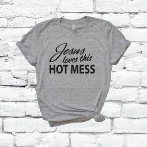 以下が含まれることがあります： グレーのTシャツに「Jesus loves this HOT MESS」という黒い文字がプリントされています。