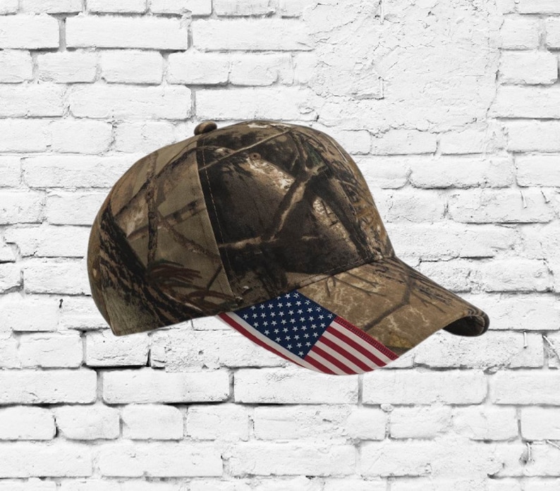 Puede incluir: Una gorra de b&eacute;isbol de camuflaje con un parche de bandera estadounidense rojo, blanco y azul en la parte delantera.