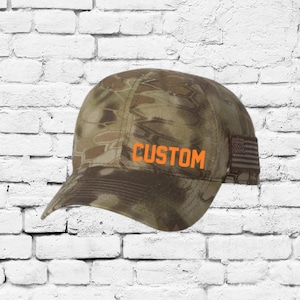 May include: A camouflage baseball cap with the word "CUSTOM" in orange block letters. The cap features a curved brim and an American flag patch on the side. The cap is designed with a brown and tan pattern.