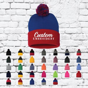Puede incluir: Una variedad de combinaciones de colores para un gorro de punto de dos tonos con un pompón en la parte superior. El gorro tiene una parte inferior roja con las palabras "Custom Embroidery" en blanco. La parte superior del gorro es azul.