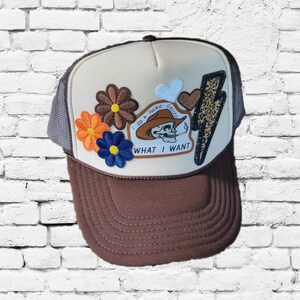 May include: A brown and beige trucker hat with a mesh back. The hat has embroidered patches of flowers, hearts, and a lightning bolt. The main patch features a skull wearing a cowboy hat and the text "I DO A THING CALLED WHAT I WANT."